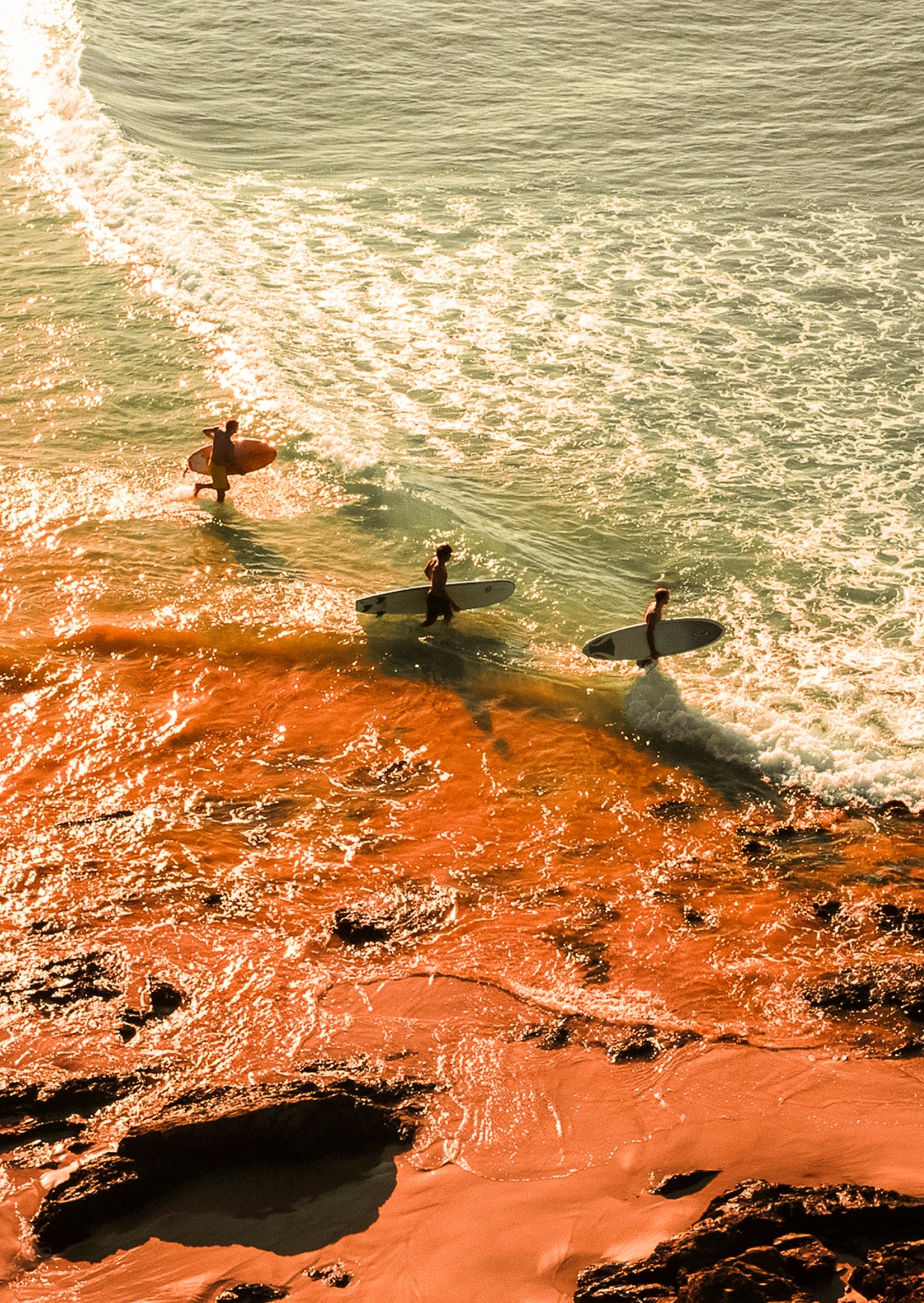 Three surfers walking along a rocky shoreline with ocean waves.