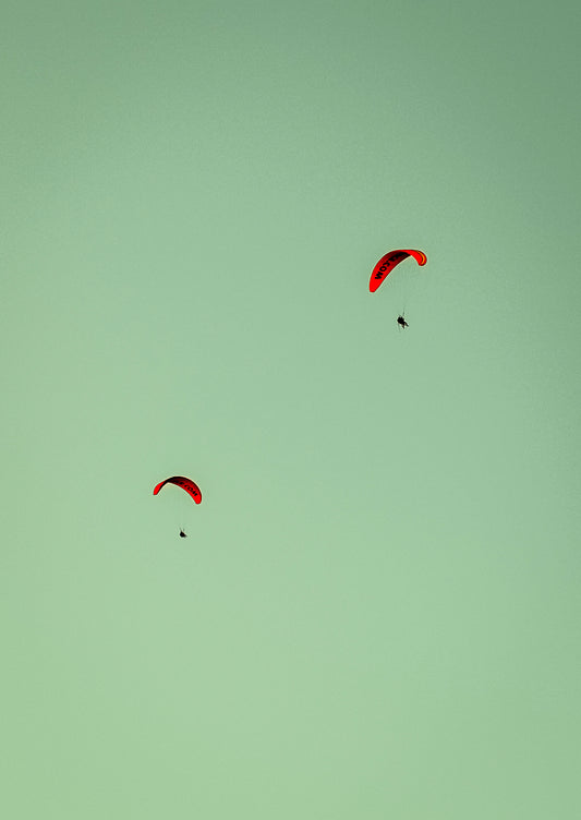 Two red paragliders flying against a light clear sky.