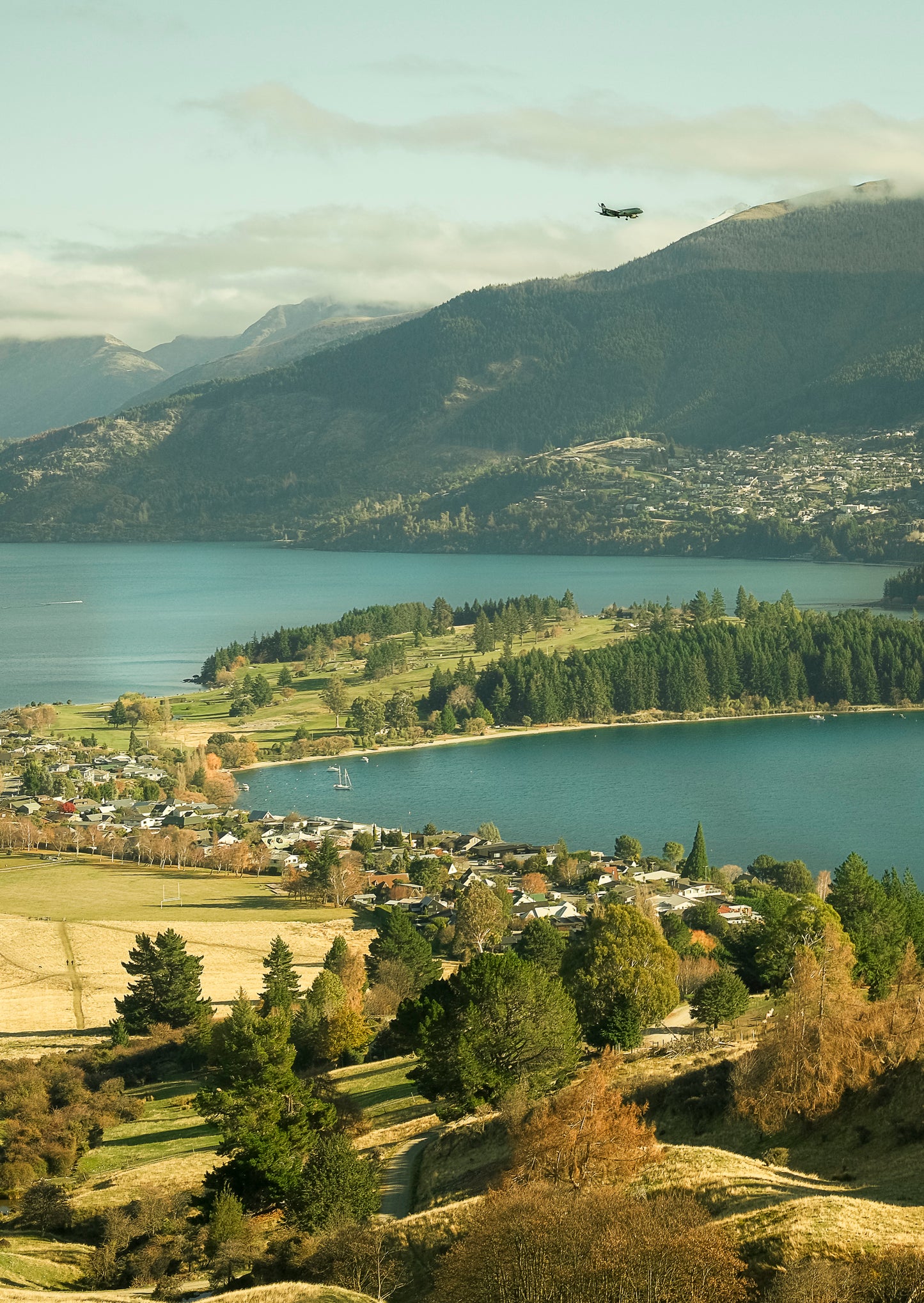Scenic view of land, a lake with mountains and a plane in the sky