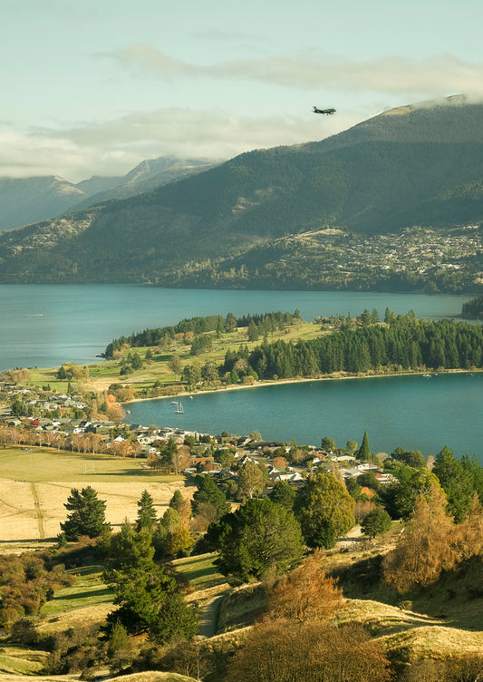 Scenic view of land, a lake with mountains and a plane in the sky