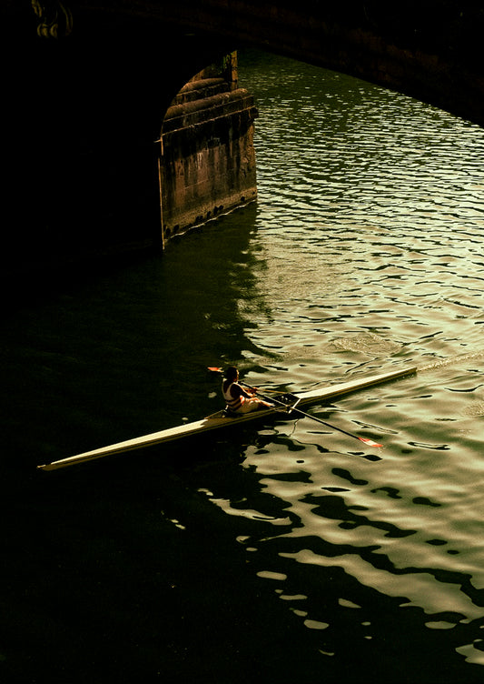 Person rowing a boat under a bridge with a dark, reflective water surface.
