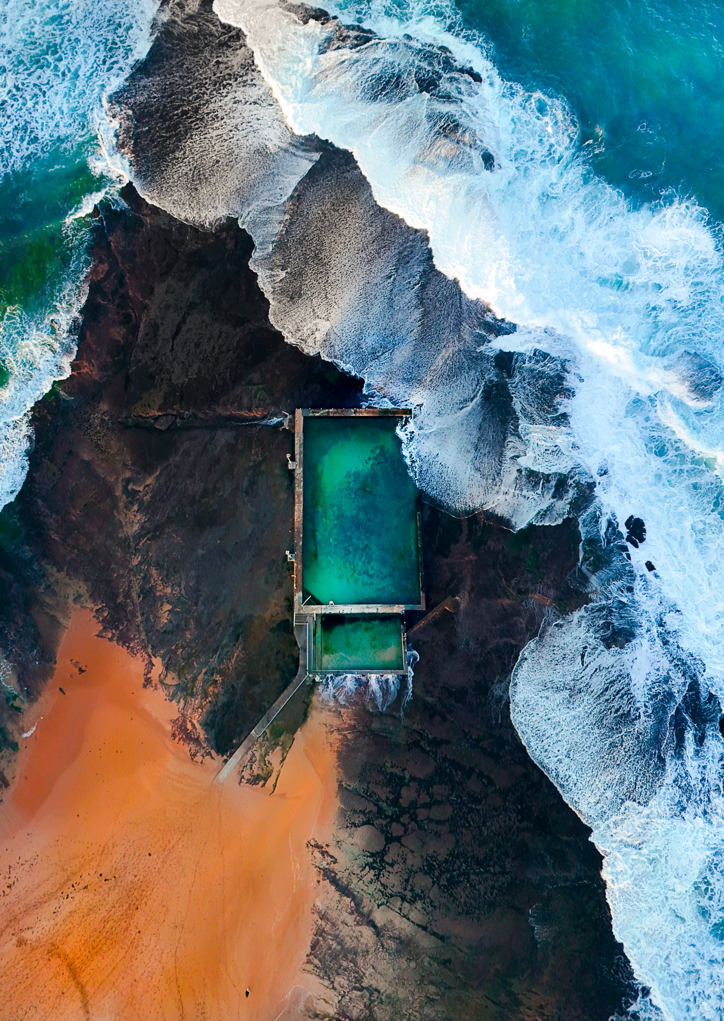 Aerial view of a pool surrounded by rugged coastline with crashing waves.