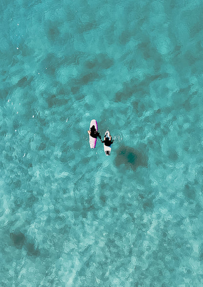 Two people sitting on surfboards on clear turquoise water from an aerial perspective.